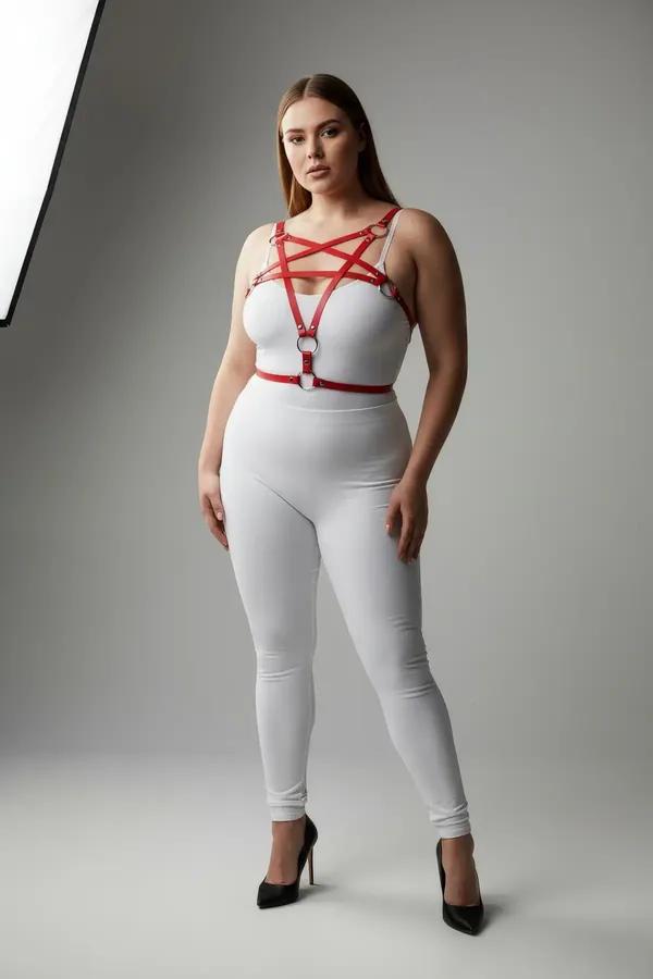 Red vegan leather starburst chest harness with silver O-rings worn over white bodysuit, plus-size model