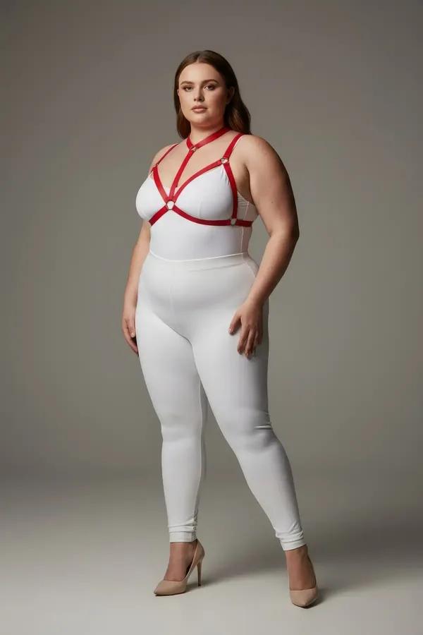 red faux leather strappy chest harness with gold O-ring accents worn over white bodysuit, plus-size model